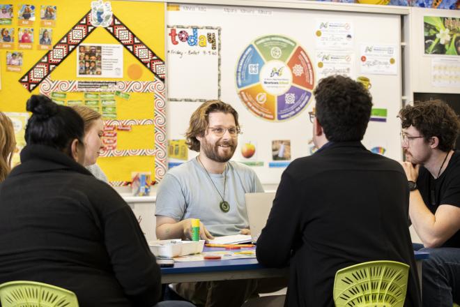 A man teacher sitting at a table talking to other teachers. He has medium-length brown hair and a beard. A man next to him is listening intently.
