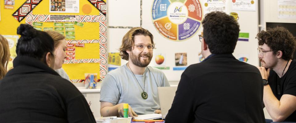 A man teacher sitting at a table talking to other teachers. He has medium-length brown hair and a beard. A man next to him is listening intently.