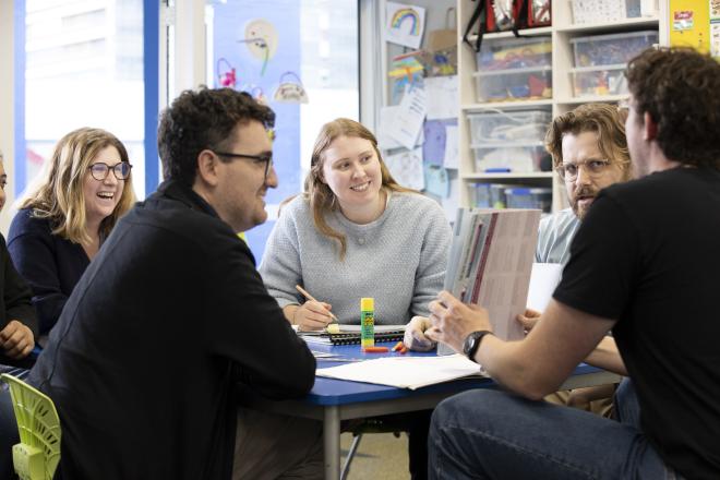 A woman teacher sitting in the centre of frame talking to a group of teachers at a table. She is smiling.