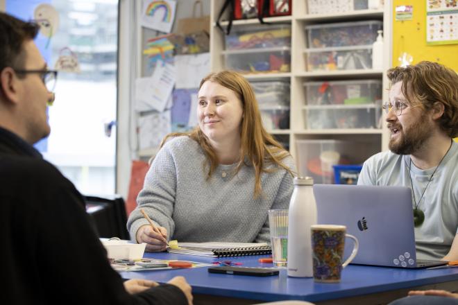 A woman teacher sitting at a table in the centre of the frame listening to other teachers speak. She has shoulder-length blonde hair. A man next to her is using a laptop. 