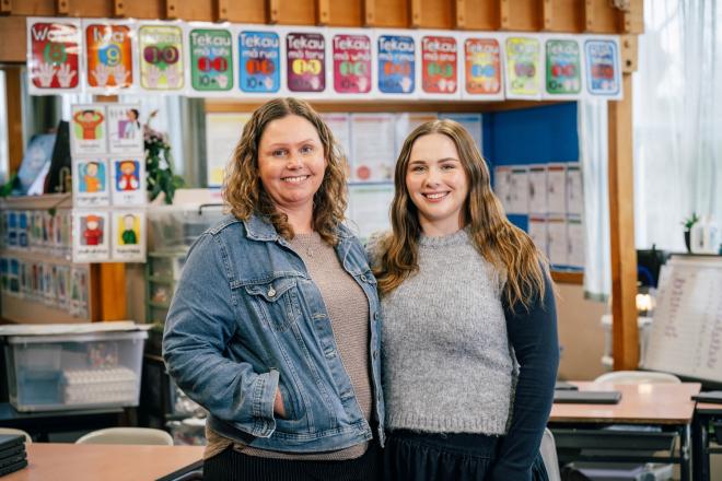 Two women stand in a classroom. They are smiling at the camera. 