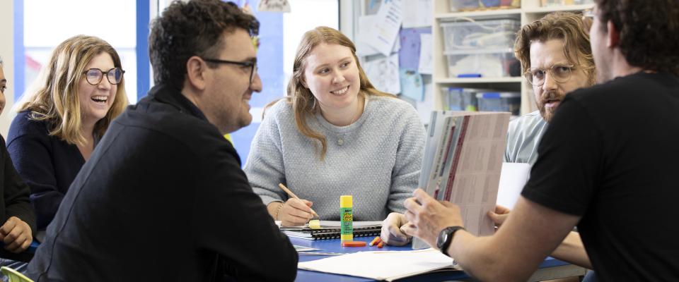 A woman teacher sitting in the centre of frame talking to a group of teachers at a table. She is smiling.