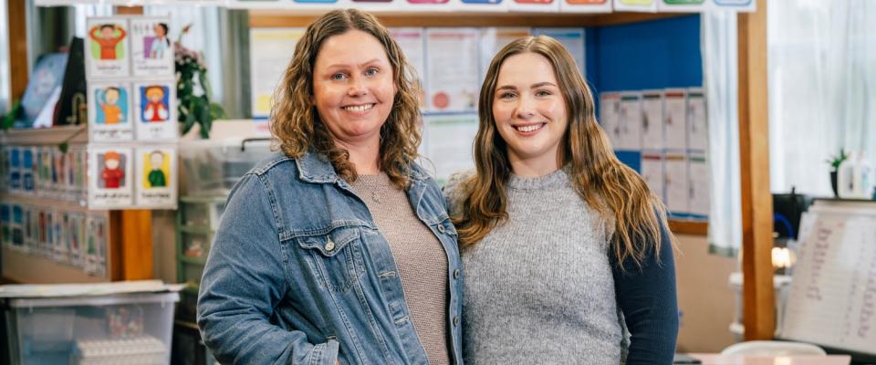 Two women stand in a classroom. They are smiling at the camera. 
